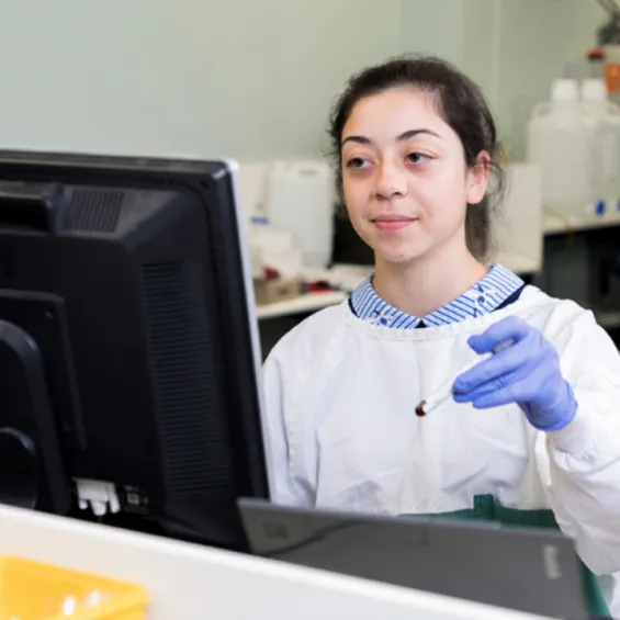 image of clinician wearing blue gloves and a lab coat holding a test tube in front of a computer monitor