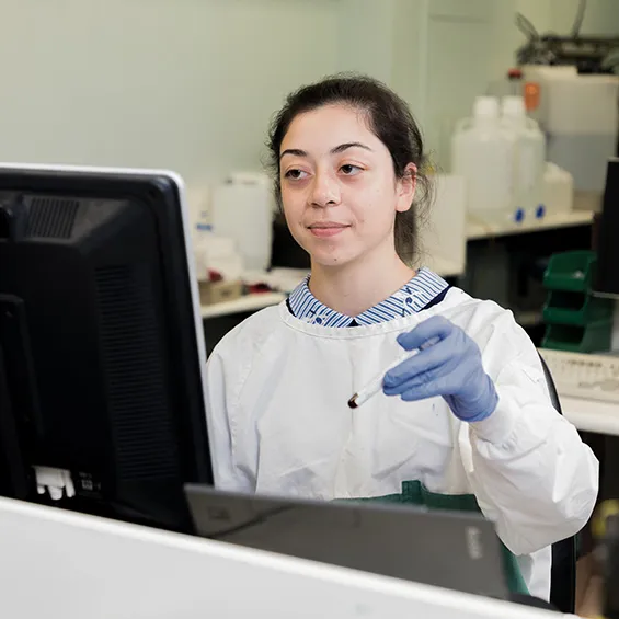 photograph of scientist wearing lab coat and blue gloves holding test tube in front of computer screen