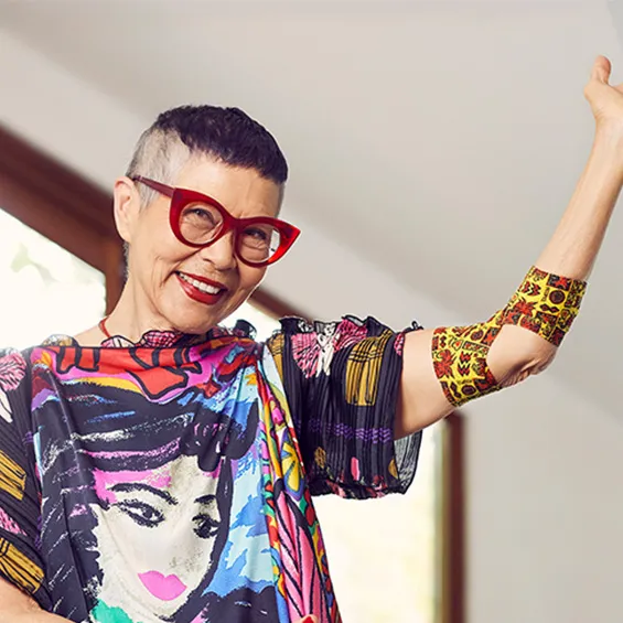 photo of jenny kee standing in a room which looks out to trees, she is wearing a colourful t-shirt and red glasses and smiling, her left arm is raised and around her elbow is a bandage with her design on it