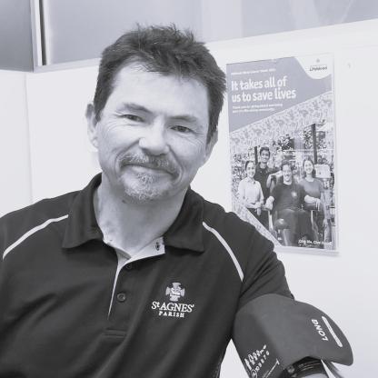A man with a blood pressure band on his arm smiling in front of a Lifeblood sign that says 'It takes all of us to save lives'