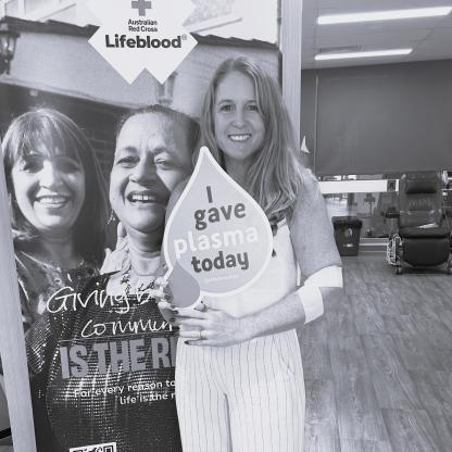 A woman standing in a donor centre in front of a Life Is the Reason sign, while holding up a donation card saying 'I gave plasma today'