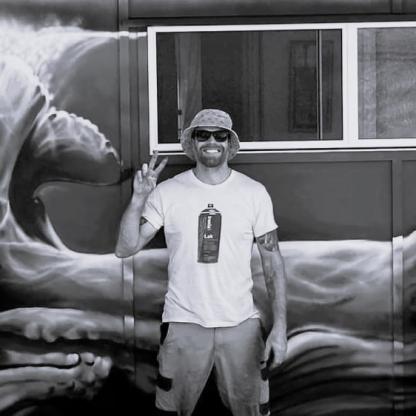 A man smiling, making the peace sign with his fingers, standing in front of a mural