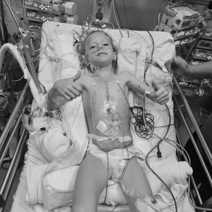 a very brave young girl in a hospital bed giving a thumbs up while hooked up to machines with wires and tubes