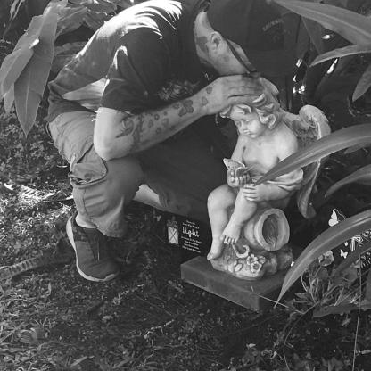 man kneeling down kissing the top of a small statue/ornament in a garden