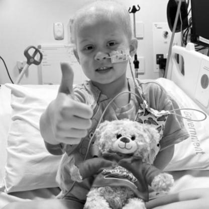 young child sitting in a hospital bed smiling and giving a thumbs up to camera