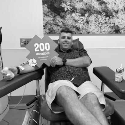man sitting in the donor chair during his donation, holding a "200 donations" sign