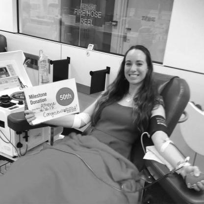 woman having her photo taken during her donation showing the needle in her arm and holding a "50th donation" sign