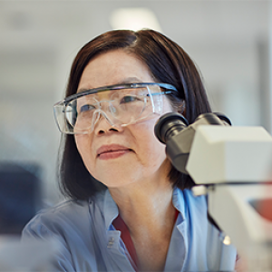 Scientist sitting in front of microscope, wearing eye protection.