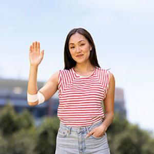 a woman is holding her hand up and smiling, on her elbow is a bandage, in the background is a stadium