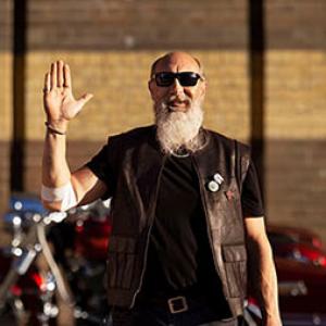 a man is holding up his hand and smiling, on his elbow is a bandage, in the background is a motorcycle
