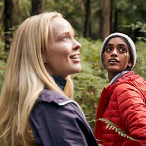 two young women are walking through a forest together, they are wearing jackets and looking up to the treetops and smiling