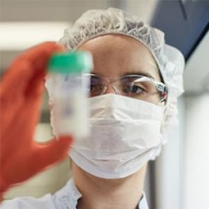 a scientist is wearing a facemask and a hairnet and is holding up a milk sample jar