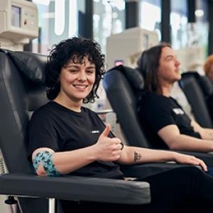 photo of a woman seated in a donor chair, smiling ang giving a thumbs up, around her right elbow is the Movember bandage