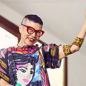 photo of jenny kee standing in a room which looks out to trees, she is wearing a colourful t-shirt and red glasses and smiling, her left arm is raised and around her elbow is a bandage with her design on it