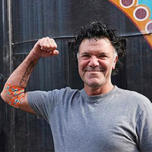 a photo of artist blak douglas smiling and holding his right arm up in a flex pose, around his elbow is the bandage he designed
