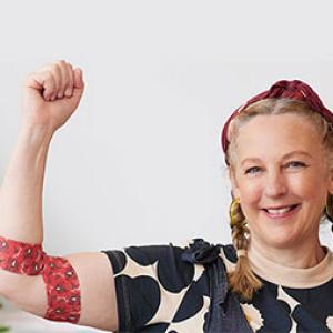 photo of artist Madeleine Stamer smiling at the camera and holding her right hand up, around her elbow is the bandage she designed