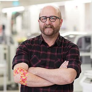 photo of artist geoff nees looking at the camera and folding his arms, around his right elbow he is wearing the bandage he designed