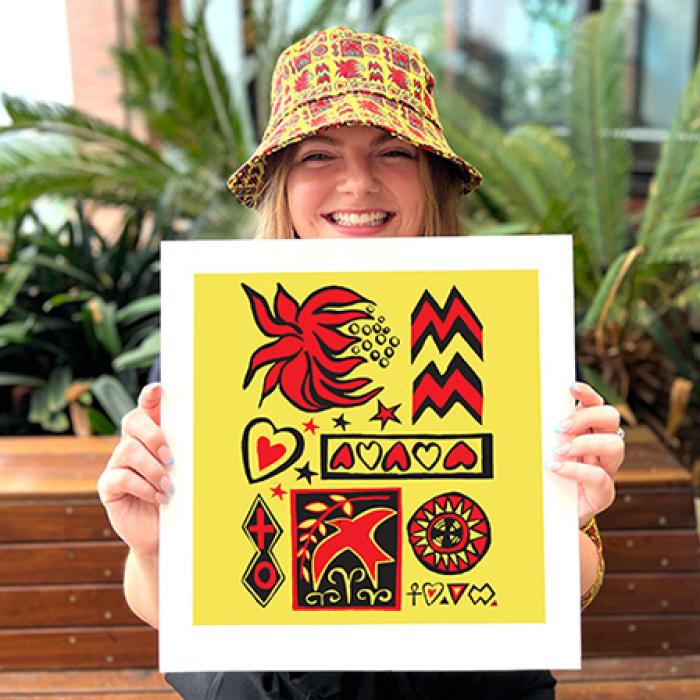 a young woman is smiling and holding up a print of Jenny Kee's design which is yellow with red and black pictures on it, she is wearing a bucket hat with the same print