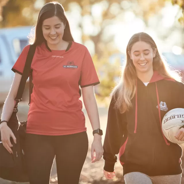 two girls are smiling and walking while wearing sportswear, they are carrying a bag and a soccer ball