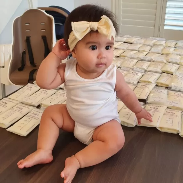 Baby with a bow tie in her hair sitting next to bags of donated breast milk