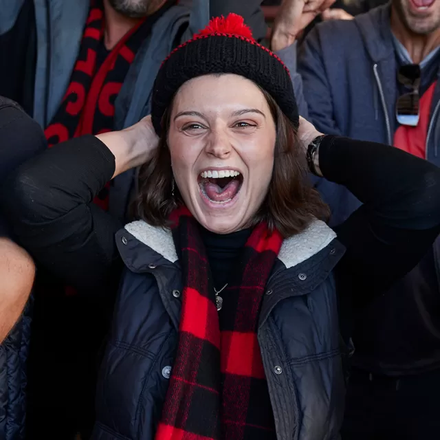 photo of a woman cheering in a crowd, she's wearing a black and red footy beanie and scarf